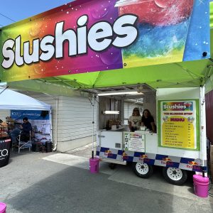 The Slushie Stop concession trailer under a lime green pop-up tent with a large rainbow Slushies banner, two women vendors smiling inside, and a menu board listing slushies and infused Red Bull
