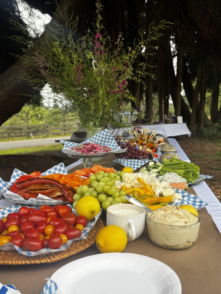 Fresh local food spread featuring fruits, vegetables, charcuterie, and appetizers at a McKinleyville community gathering surrounded by redwood trees.