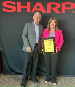 Two people stand in front of a SHARP backdrop while holding a SHARP HK award presented to Coastal Business Systems.